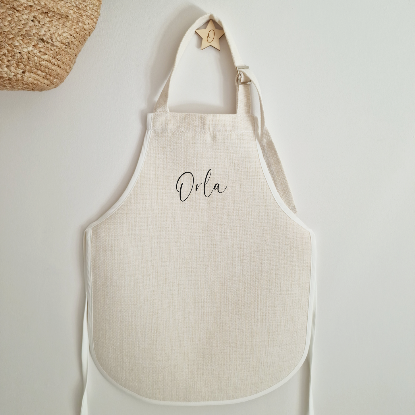 Personalised off white apron with name in a minimalist script font, hanging on a wall