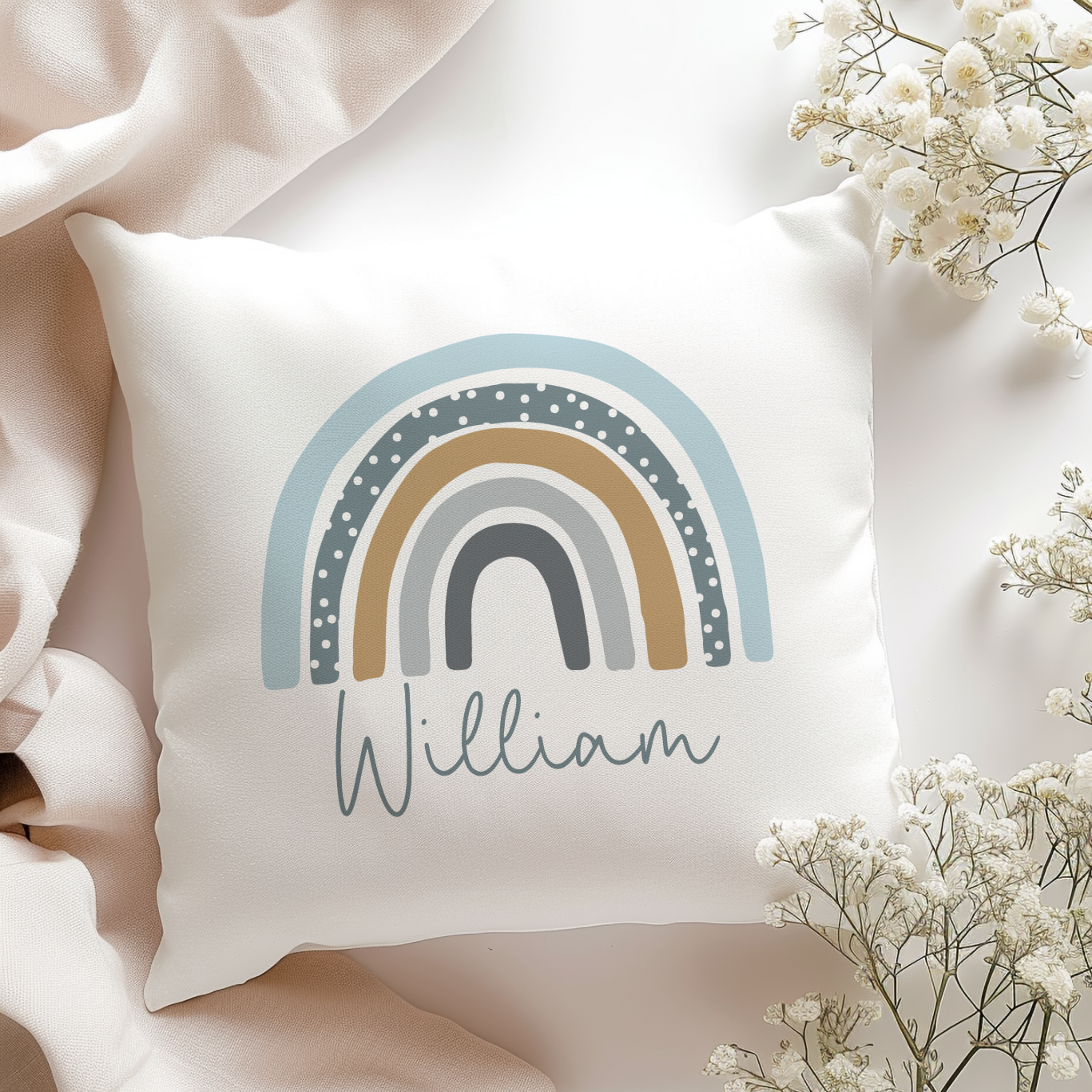 White pillow with blue rainbow design and 'William' text, surrounded by light-colored fabric and small white flowers.