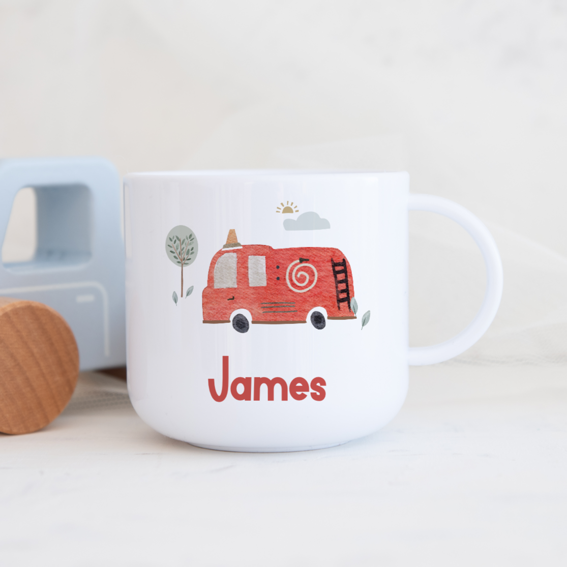 Children's mug with a red truck design and 'James' text, next to a toy truck on a light background