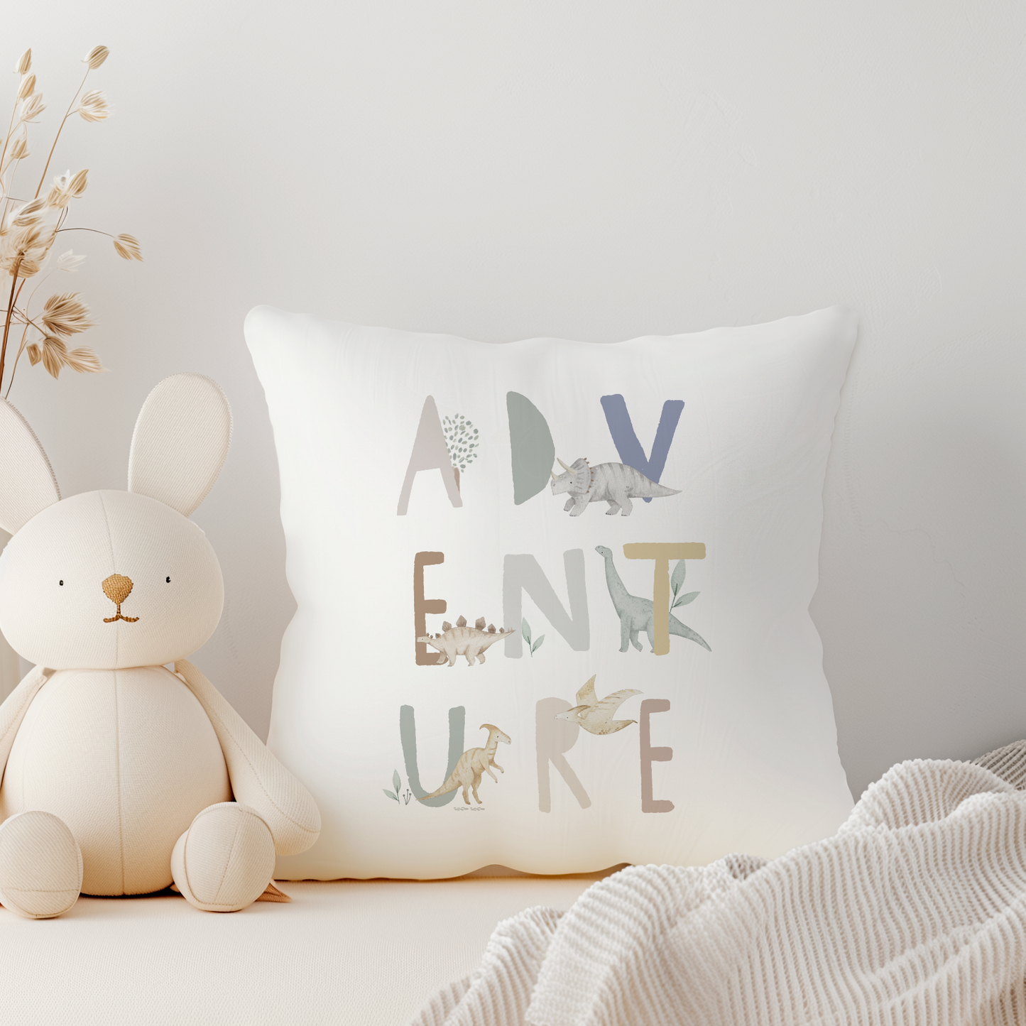 White canvas cushion with dinosaur adventure, in muted and blue and green tones with dinosaur illustration around letters