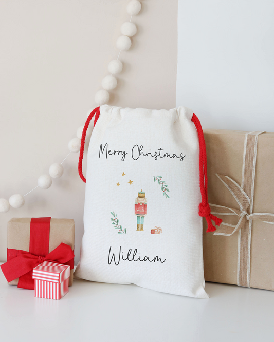 White gift sack with 'Merry Christmas' and a nutcracker design, surrounded by small gifts on a light background.