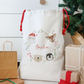 White drawstring bag with festive designs and 'Amelia' text, surrounded by Christmas presents and decorations.