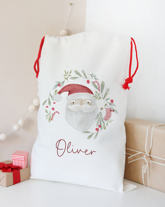 White drawstring bag with Santa Claus design and 'Oliver' text, surrounded by Christmas decorations.