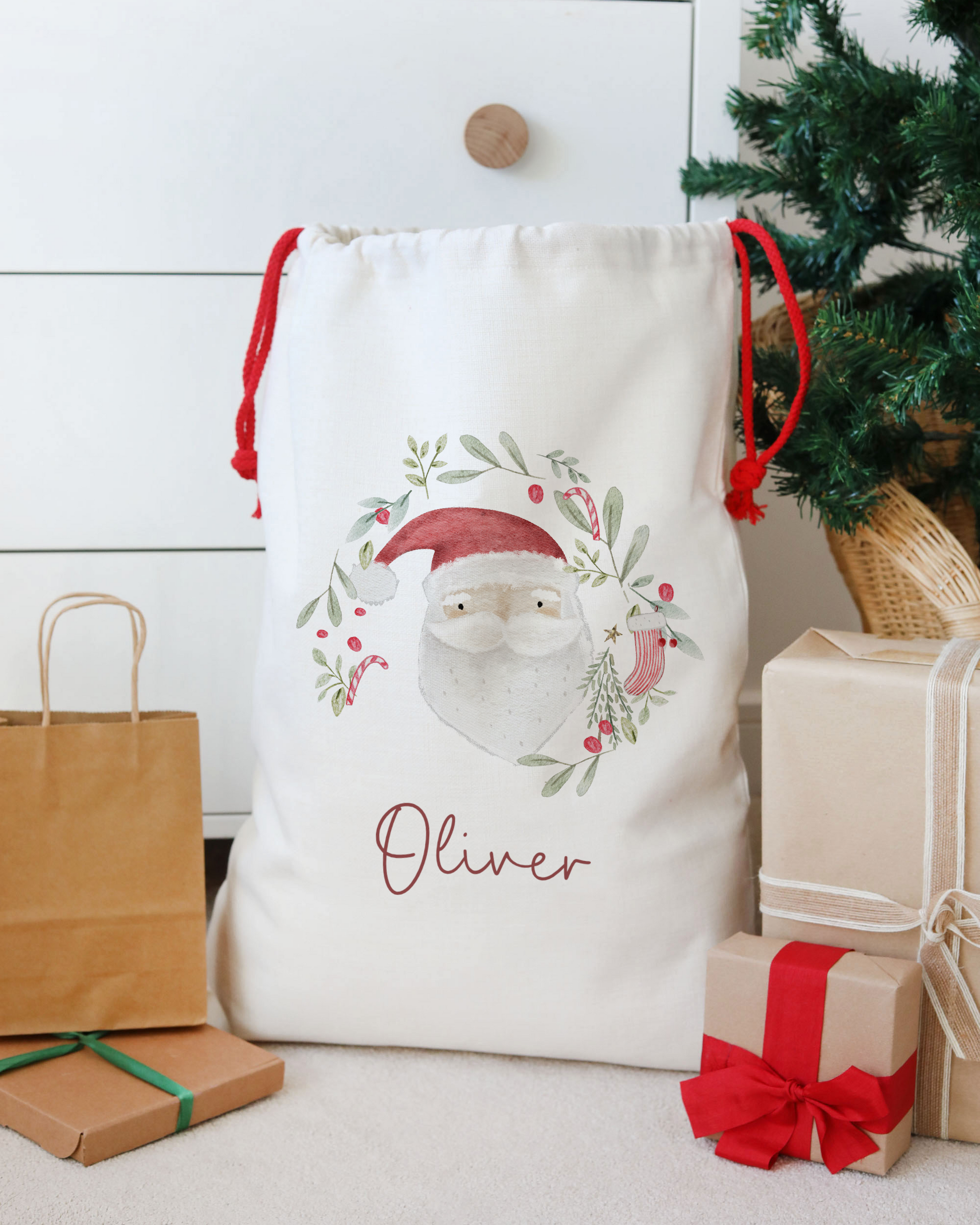 White gift bag with Santa Claus design and 'Oliver' text, surrounded by Christmas presents and decorations.