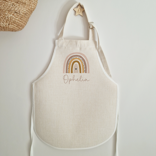 Child's off white linen-like apron. Personalised with name featuring a boho rainbow in burnt orange and pink tones.