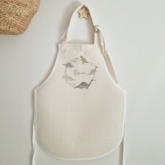 Childs off white linen look apron with dinosaur wreath illustration. Personalised with  name in dark grey script font.