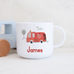 Children's mug with a red truck design and 'James' text, next to a toy truck on a light background