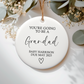Round ceramic ornament with text 'You're going to be a Grandad Baby Harrison Due May 2025' on a white background with greenery.