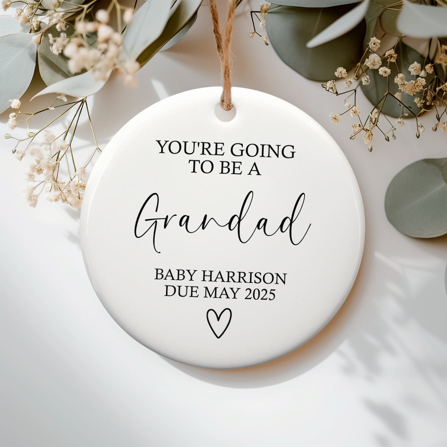 Round ceramic ornament with text 'You're going to be a Grandad Baby Harrison Due May 2025' on a white background with greenery.