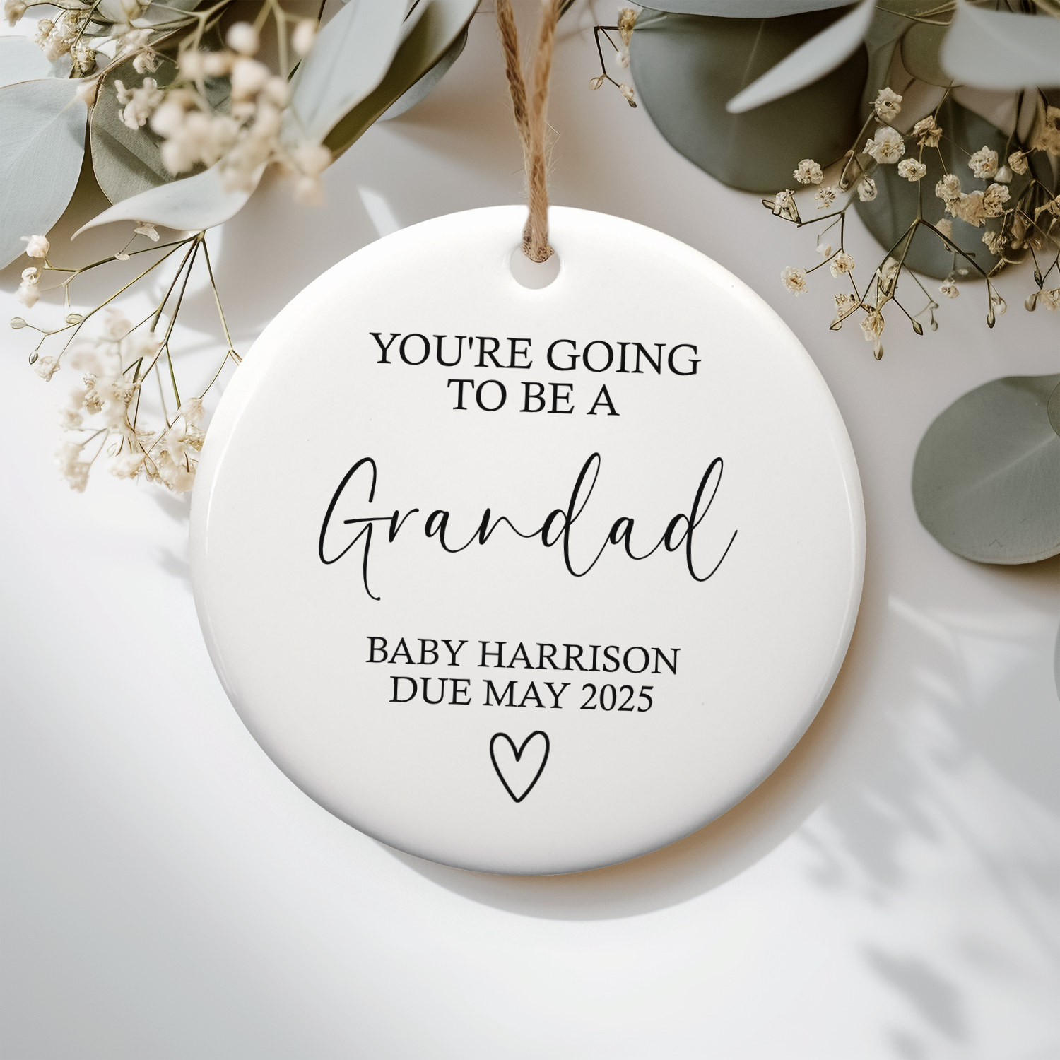 Round ceramic ornament with text 'You're going to be a Grandad Baby Harrison Due May 2025' on a white background with greenery.
