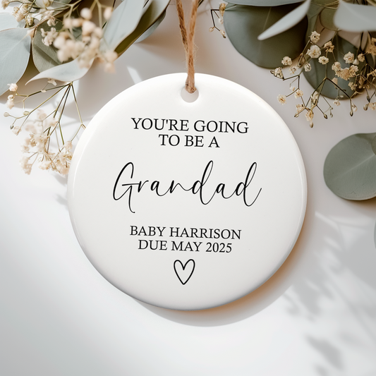 Round ceramic ornament with text 'You're going to be a Grandad Baby Harrison Due May 2025' on a white background with greenery.