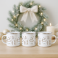 Three white mugs with decorative patterns on a wooden surface in front of a Christmas wreath.