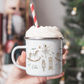 Hand holding a mug of hot chocolate with whipped cream and a red and white straw, Christmas tree in the background. Mug has traditional nutcracker themed illustrations.