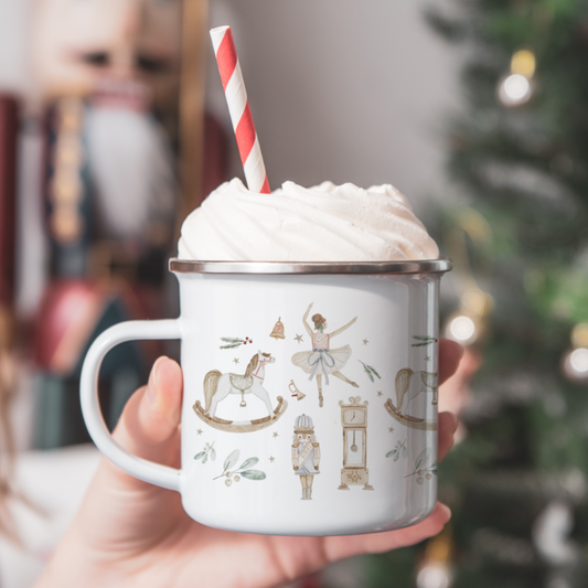 Hand holding a mug of hot chocolate with whipped cream and a red and white straw, Christmas tree in the background. Mug has traditional nutcracker themed illustrations.